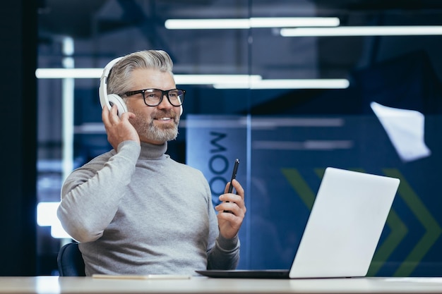 Uomo dai capelli grigi sorridente anziano seduto alla scrivania in ufficio che indossa le cuffie davanti ai colloqui del laptop