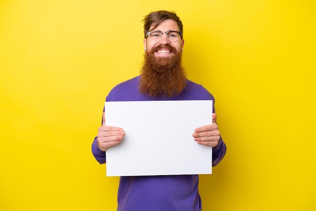 Uomo dai capelli rossi con la barba isolato su sfondo giallo che tiene un cartello vuoto con un'espressione felice