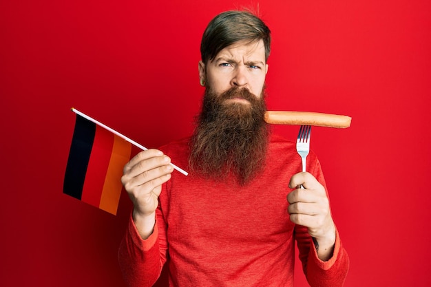 uomo dai capelli rossi con la barba lunga che tiene la forchetta con salsiccia di maiale e bandiera tedesca scettico e nervoso accigliato sconvolto a causa del problema persona negativa