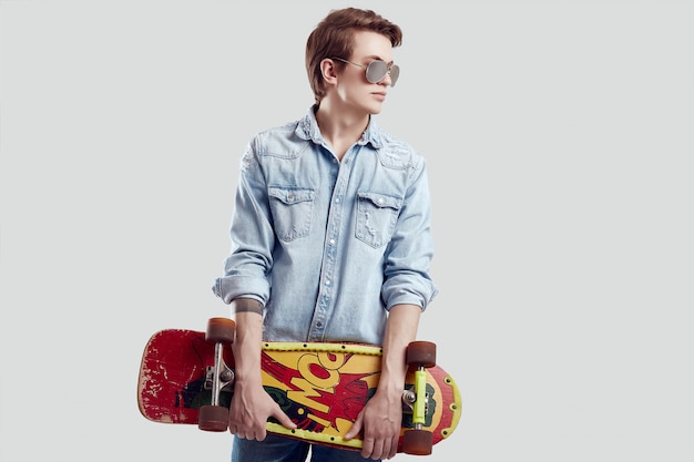 Uomo dei pantaloni a vita bassa in giacca da sole e jeans che posa con lo skateboard