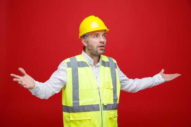 Uomo del costruttore in uniforme da costruzione e casco di sicurezza che guarda da parte confuso allargando le braccia ai lati senza risposta in piedi su sfondo rosso