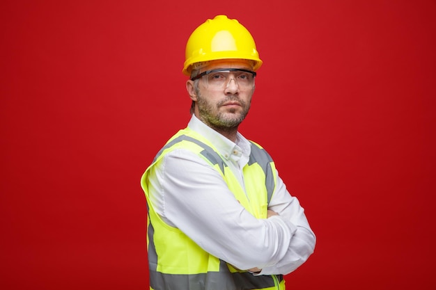 Uomo del costruttore in uniforme da costruzione e casco di sicurezza che indossa occhiali di sicurezza guardando la fotocamera con la faccia seria con le braccia incrociate sul petto in piedi su sfondo rosa