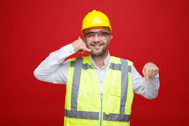 Uomo del costruttore in uniforme da costruzione e casco di sicurezza che indossa occhiali di sicurezza guardando la fotocamera sorridendo allegramente facendomi chiamare gesto puntando con il dito indice in basso in piedi su sfondo rosso