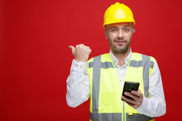 Uomo del costruttore in uniforme da costruzione e messaggio di testo del casco di sicurezza utilizzando lo smartphone che punta con il pollice di lato guardando sicuro in piedi su sfondo rosso