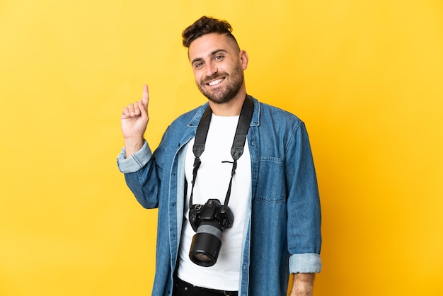 Uomo del fotografo isolato sulla parete gialla che mostra e che solleva un dito in segno del meglio