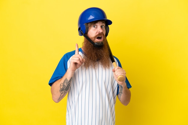 Uomo del giocatore di baseball della testarossa con casco e pipistrello isolato su sfondo giallo pensando a un'idea che punta il dito verso l'alto