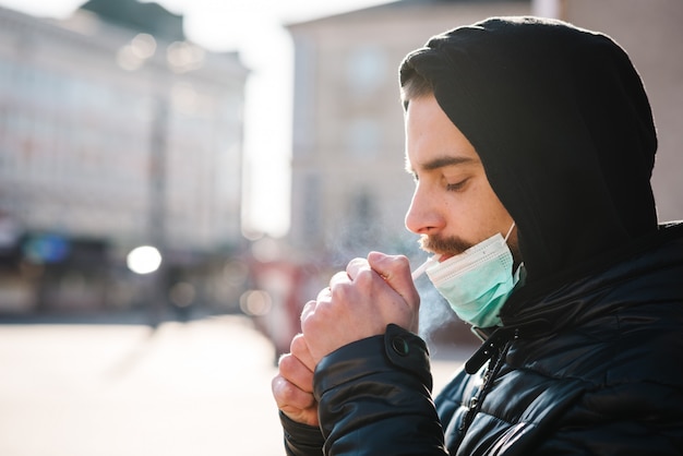 Uomo del primo piano con la maschera durante la pandemia COVID-19 che fuma una sigaretta alla via.