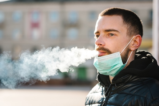 Uomo del primo piano con la maschera durante la pandemia COVID-19 che fuma una sigaretta alla via.