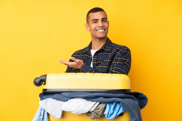 Uomo del viaggiatore con una valigia piena di vestiti sopra la parete gialla isolata che estende le mani al lato per invitare a venire