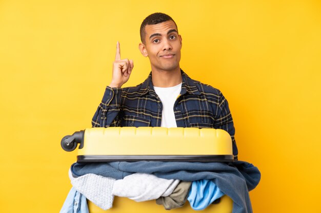 Uomo del viaggiatore con una valigia piena di vestiti sopra la parete gialla isolata che indica con il dito indice una grande idea