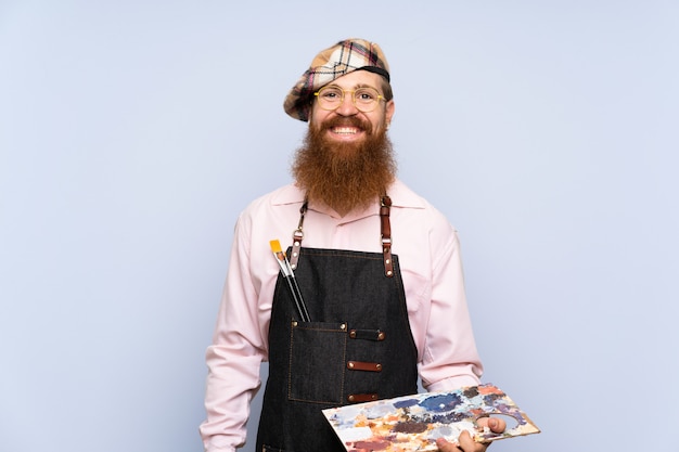 Uomo dell'artista della testarossa con la barba lunga che tiene una tavolozza sopra fondo blu isolato che sorride molto