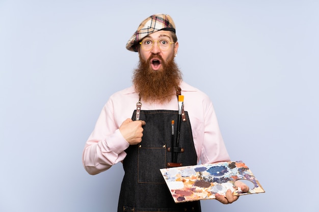 Uomo dell'artista della testarossa con la barba lunga che tiene una tavolozza sopra fondo blu isolato con espressione facciale di sorpresa