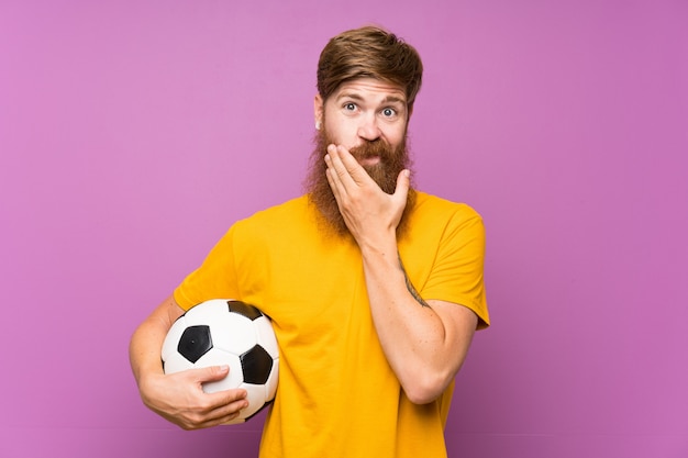 Uomo della testarossa con la barba lunga che tiene un pallone da calcio sopra la parete porpora isolata che pensa un'idea