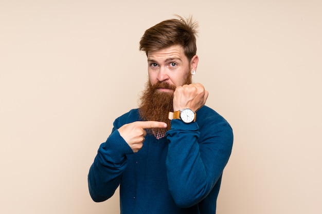 Uomo della testarossa con la barba lunga sopra fondo isolato che mostra l'orologio della mano con l'espressione seria