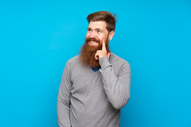 Uomo della testarossa con la barba lunga sopra il blu isolato che pensa un'idea mentre osservando in su