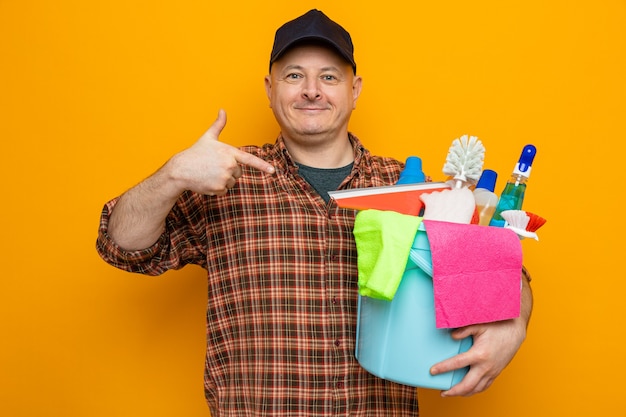 Uomo delle pulizie in camicia a quadri e cappello che tiene il secchio con strumenti per la pulizia che puntano con il dito indice verso di loro guardando la telecamera sorridente fiducioso in piedi su sfondo arancione