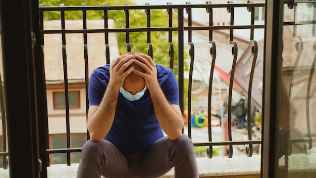 Uomo depresso con maschera usa e getta seduto in balcone durante l'epidemia di coronavirus.