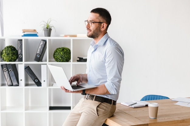 Uomo di affari di profilo che indossa una camicia bianca seduta sul tavolo in ufficio e guardando avanti mentre si utilizza il computer portatile d'argento per il lavoro