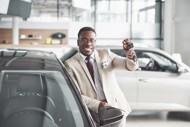 Uomo di colore bello in concessionaria sta abbracciando la sua nuova auto e sorridente