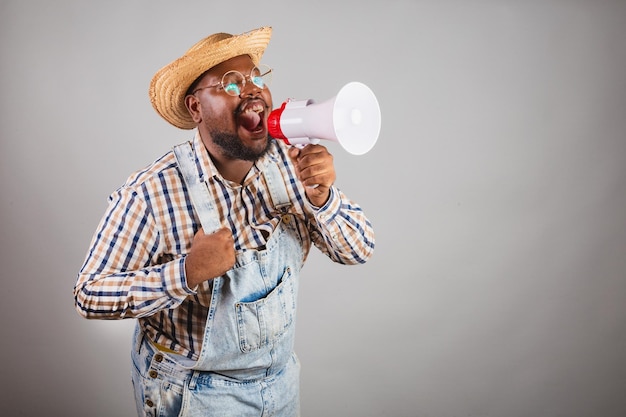 Uomo di colore brasiliano che indossa abiti da campagna dalla festa junina festa de sao joao arraia usando il megafono che grida la promozione annunciando lo sconto pubblicitario