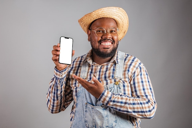 Uomo di colore brasiliano che indossa abiti da campagna della festa junina festa de sao joao arraia con in mano uno smartphone che mostra app mobili con schermo vuoto