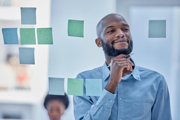 Uomo di colore che scrive e pensa nel programma di pianificazione su una lavagna di vetro per attività di brainstorming in ufficio Maschio africano che si chiede nel pensiero per l'idea del piano di progetto o una nota adesiva per la strategia aziendale