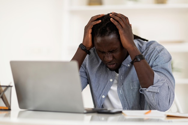 Uomo di colore depresso che soffre di mal di testa mentre si lavora con il computer portatile in ufficio a casa