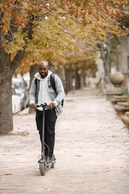 Uomo di colore in piedi su una strada con scooter elettrico. Uomo che indossa una felpa con cappuccio bianca e jeans neri