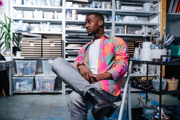 Uomo di colore rilassato in camicia fantasia quadrata colorata seduto su una sedia in un'officina. Tutti i tipi di contenitori sugli scaffali in sottofondo.