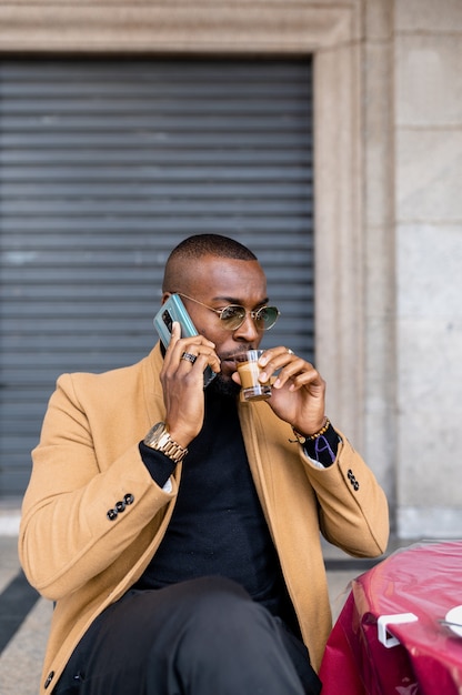 Uomo di colore seduto in un bar a tavola a bere caffè mentre parla al telefono. Colpo verticale.