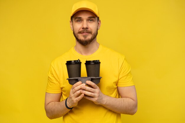 Uomo di consegna allegro in uniforme gialla che sorride e che tiene le tazze di caffè da asporto isolate