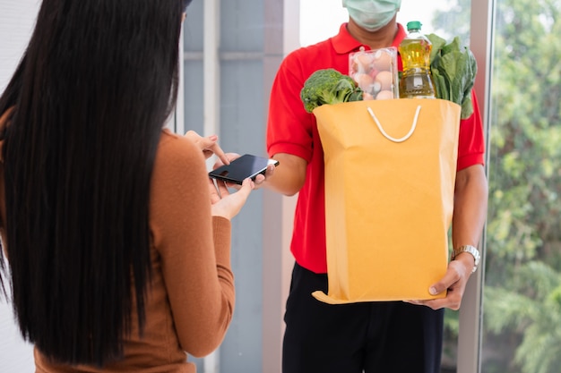 Uomo di consegna asiatico che tiene un sacchetto di cibo fresco per dare ai clienti e che tiene smartphone per ricevere pagamenti a casa