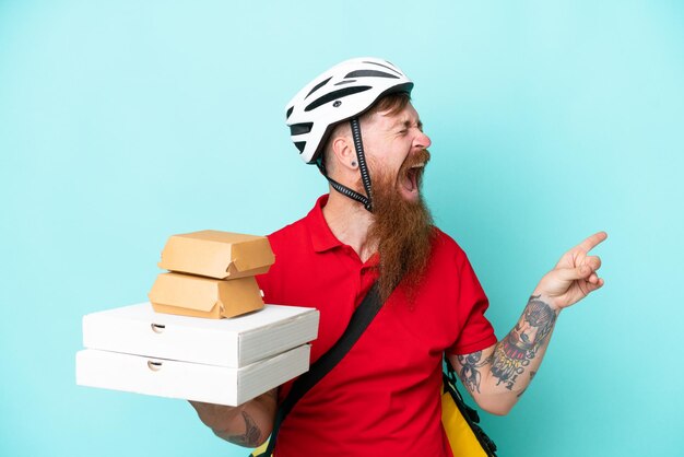 Uomo di consegna che tiene pizze e hamburger isolati su sfondo blu che punta il dito di lato e presenta un prodotto