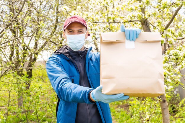 Uomo di consegna che tiene un sacchetto di carta con cibo su sfondo bianco, fattorino di cibo in maschera protettiva e guanti protettivi.