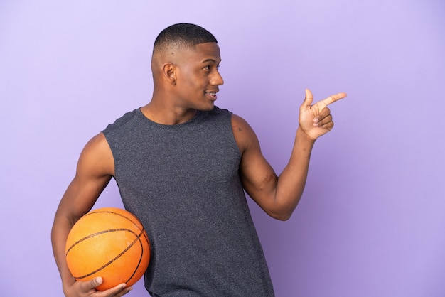 Uomo di giovane giocatore di basket latino isolato sul muro viola che punta il dito a lato e presenta un prodotto