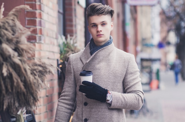 Uomo di hipster in cappotto di autunno che beve caffè sulla strada della città