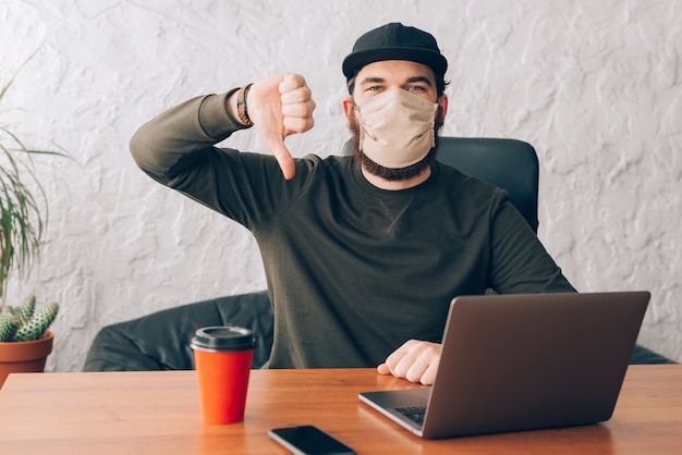 Uomo di impiegato che sta davanti al computer portatile con l'anti maschera del virus e che mostra i pollici giù