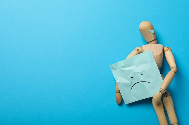 Uomo di legno con carta con un'emoji triste su sfondo blu spazio per il testo