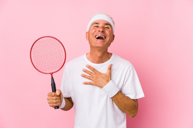 Uomo di mezza età che gioca a badminton isolato ride ad alta voce tenendo la mano sul petto.