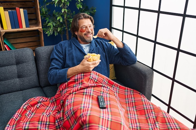 Uomo di mezza età che indossa bende sotto gli occhi guardando film che mangia patate fritte a casa
