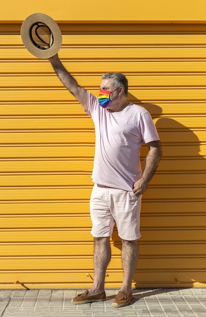 Uomo di mezza età che indossa un cappello, occhiali da sole e una maschera protettiva color arcobaleno. LGTB su sfondo giallo. Concetto Covid-19