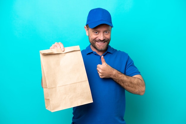 Uomo di mezza età che prende un sacchetto di cibo da asporto isolato su sfondo blu dando un gesto di pollice in alto