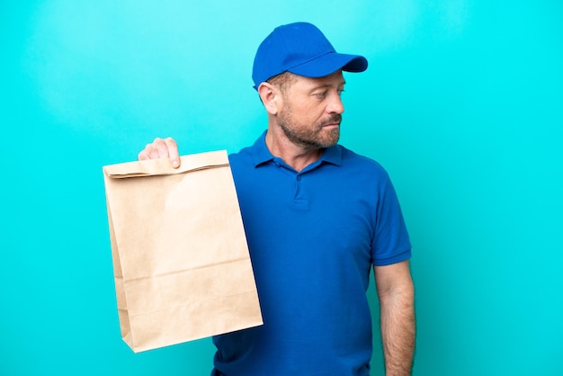 Uomo di mezza età che prende un sacchetto di cibo da asporto isolato su sfondo blu guardando di lato