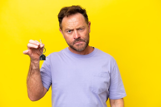 Uomo di mezza età che tiene le chiavi di casa isolate su sfondo giallo con espressione triste