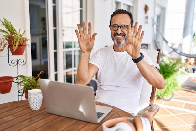 Uomo di mezza età che utilizza il computer portatile a casa mostrando e indicando con le dita numero nove mentre sorride fiducioso e felice.