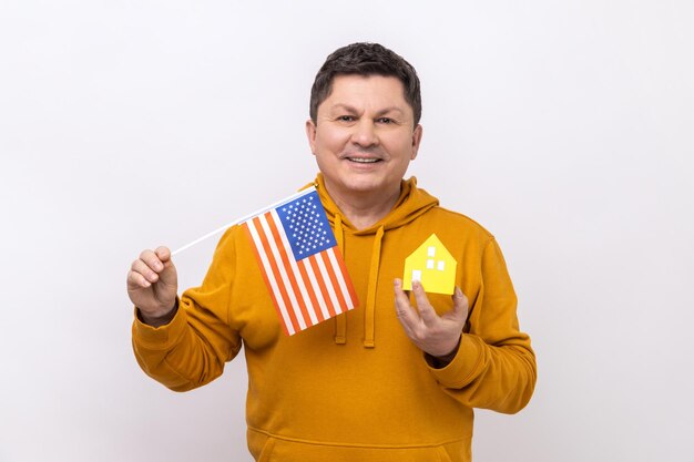 Uomo di mezza età con bandiera americana e casa di carta che sogna di acquistare un alloggio negli Stati Uniti