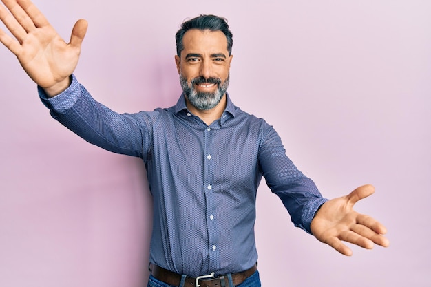 Uomo di mezza età con barba e capelli grigi che indossa abiti da lavoro che guarda la telecamera sorridendo con le braccia aperte per un abbraccio espressione allegra abbracciando la felicità