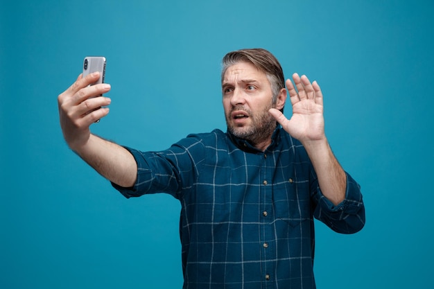 Uomo di mezza età con capelli grigi in camicia di colore scuro che tiene smartphone con videochiamata che sembra preoccupato agitando con la mano in piedi su sfondo blu