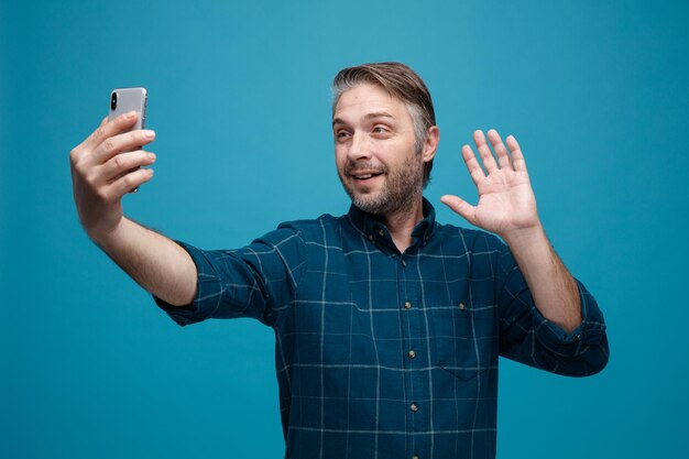 Uomo di mezza età con capelli grigi in camicia di colore scuro che tiene smartphone con videochiamata sventolando con gesto di saluto della mano sorridente amichevole in piedi su sfondo blu