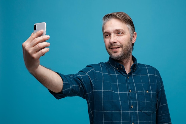 Uomo di mezza età con i capelli grigi in camicia di colore scuro che fa selfie utilizzando lo smartphone sorridente allegramente felice e positivo in piedi su sfondo blu